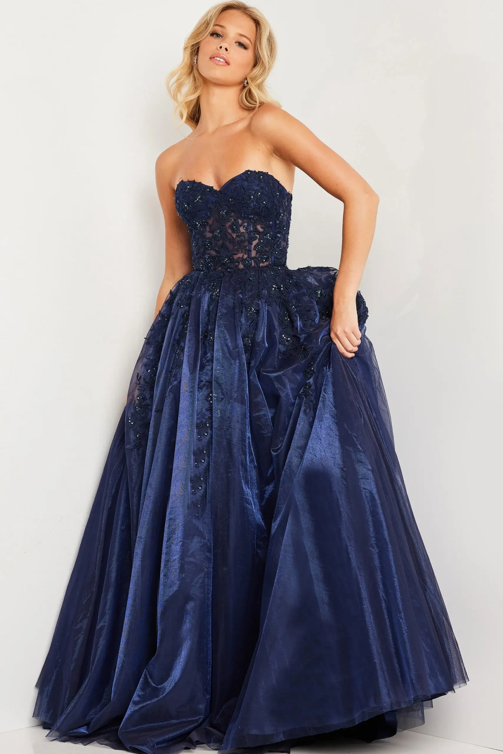 A woman in a strapless, navy blue ball gown adorned with floral lace detail stands confidently against a neutral background, exuding elegance and poise.