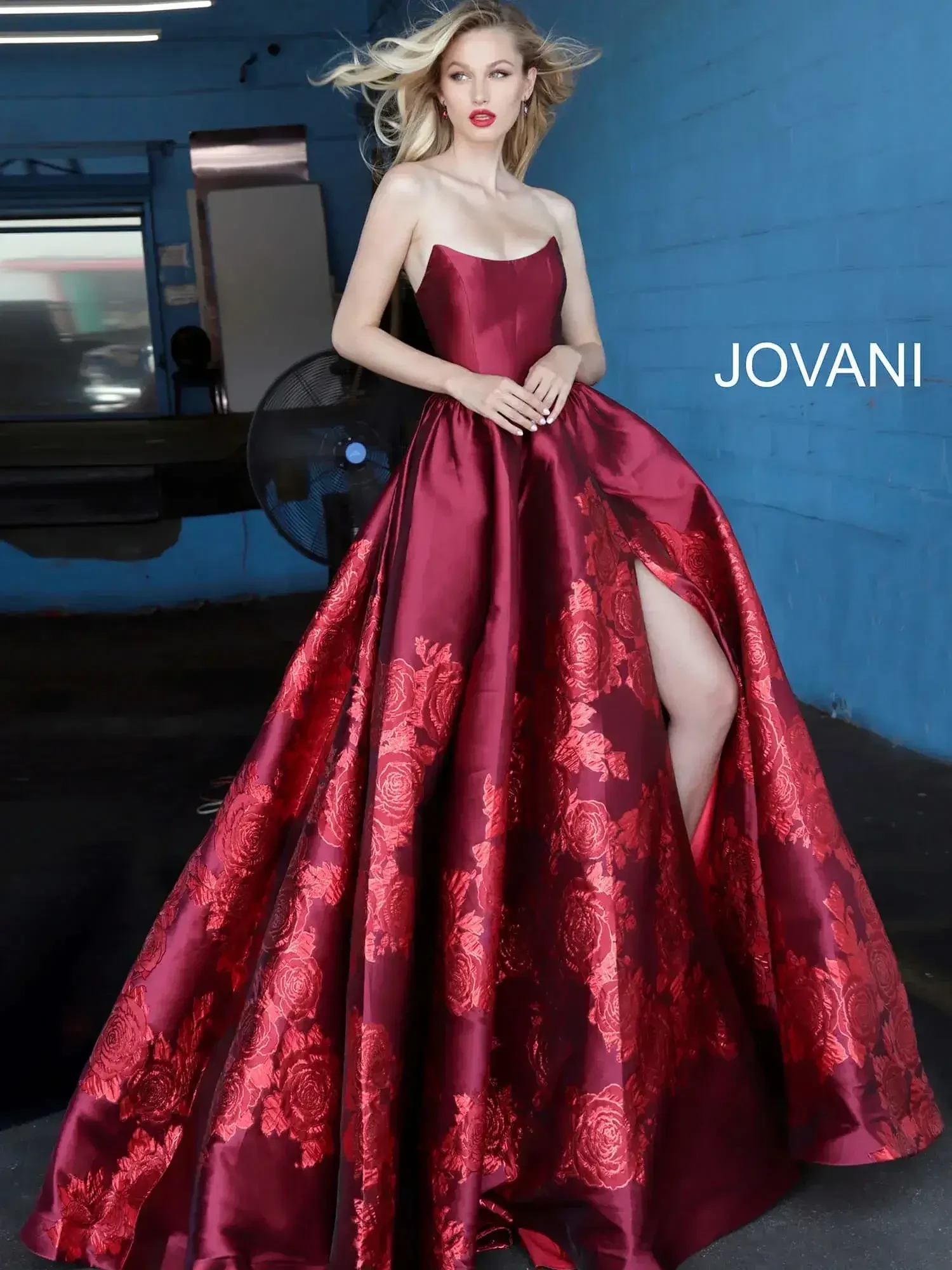 A woman in a strapless, burgundy ball gown with floral patterns poses confidently indoors. Her dress has a high slit, and her expression is elegant and poised.