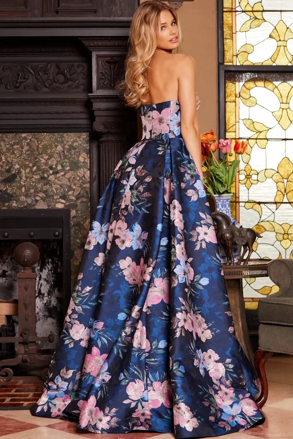 A woman in a floral patterned gown stands by a fireplace, with a stained glass window behind her.