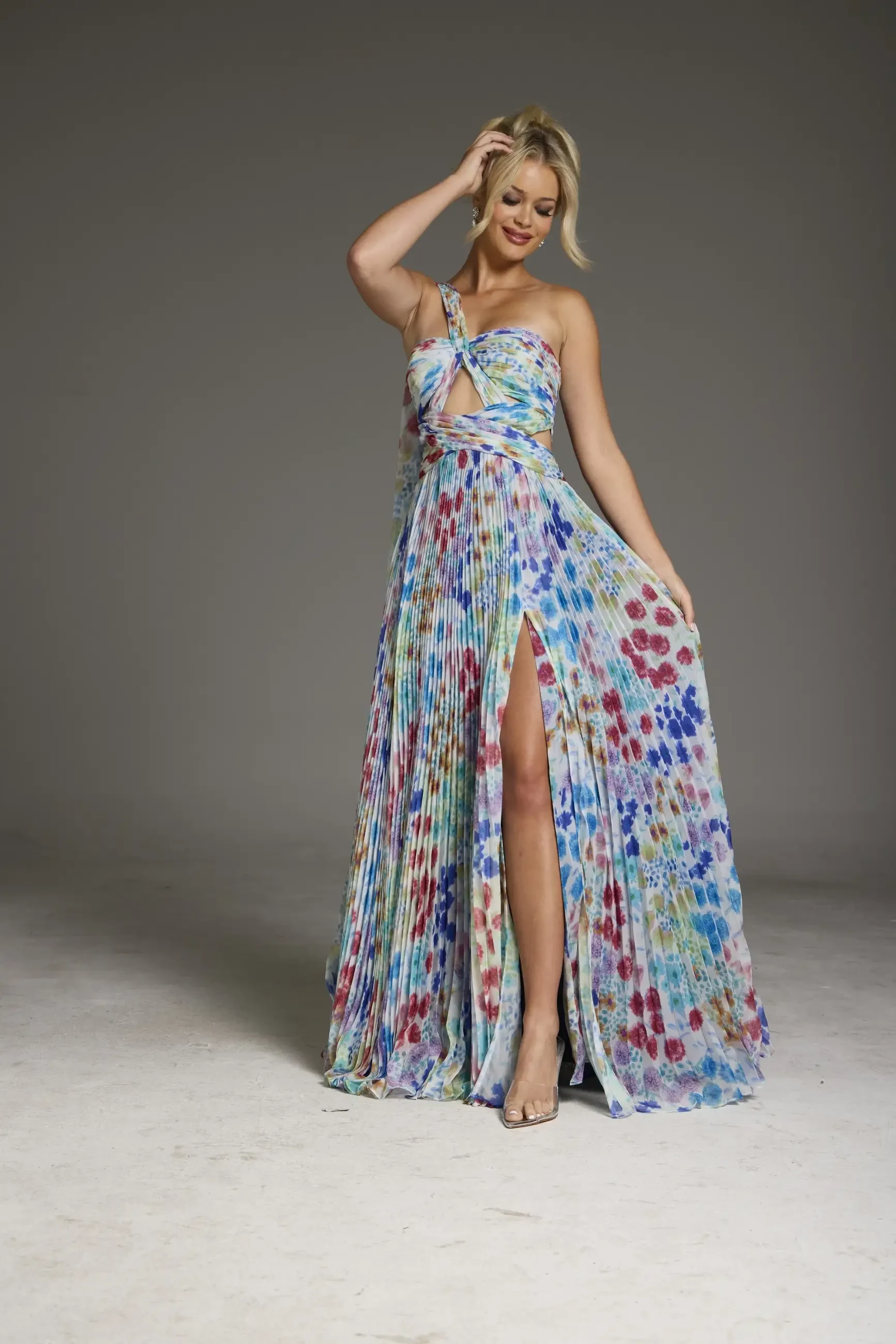 Woman in a colorful, floral maxi dress poses with one hand in her hair. The dress features a thigh-high slit and a cut-out at the chest, set in a studio.