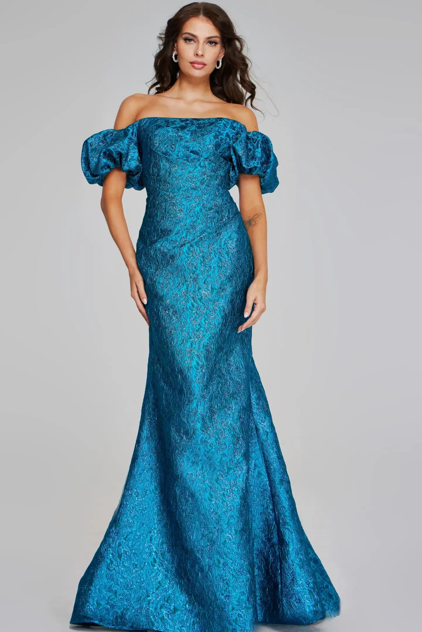 Model is wearing a teal off-the-shoulder mermaid gown with puffy short sleeves, fitted bodice and hips, a flowing skirt, and a metallic floral pattern.