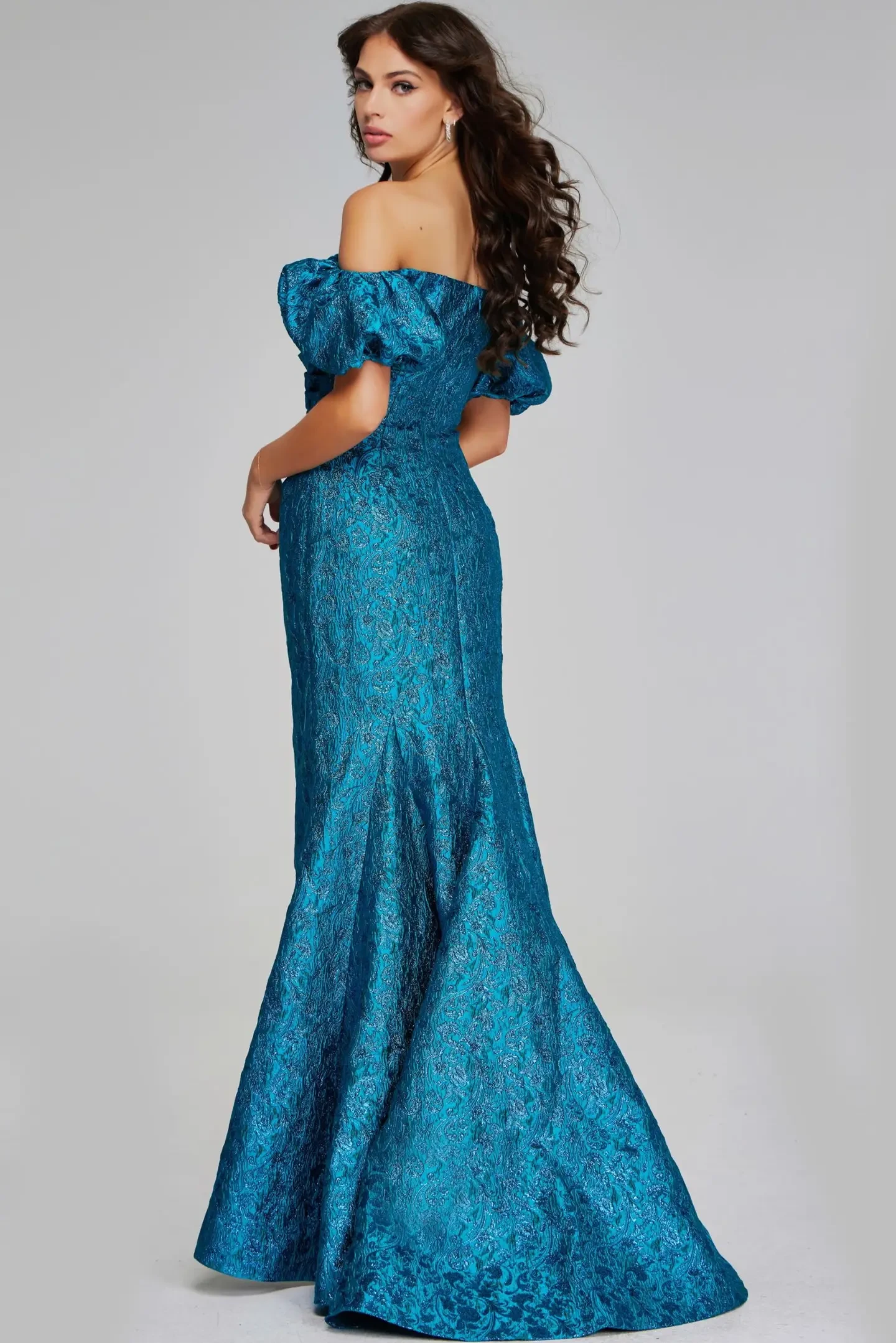 Model is wearing a teal off-the-shoulder mermaid gown with puffy short sleeves, fitted bodice and hips, a flowing skirt, and a metallic floral pattern.