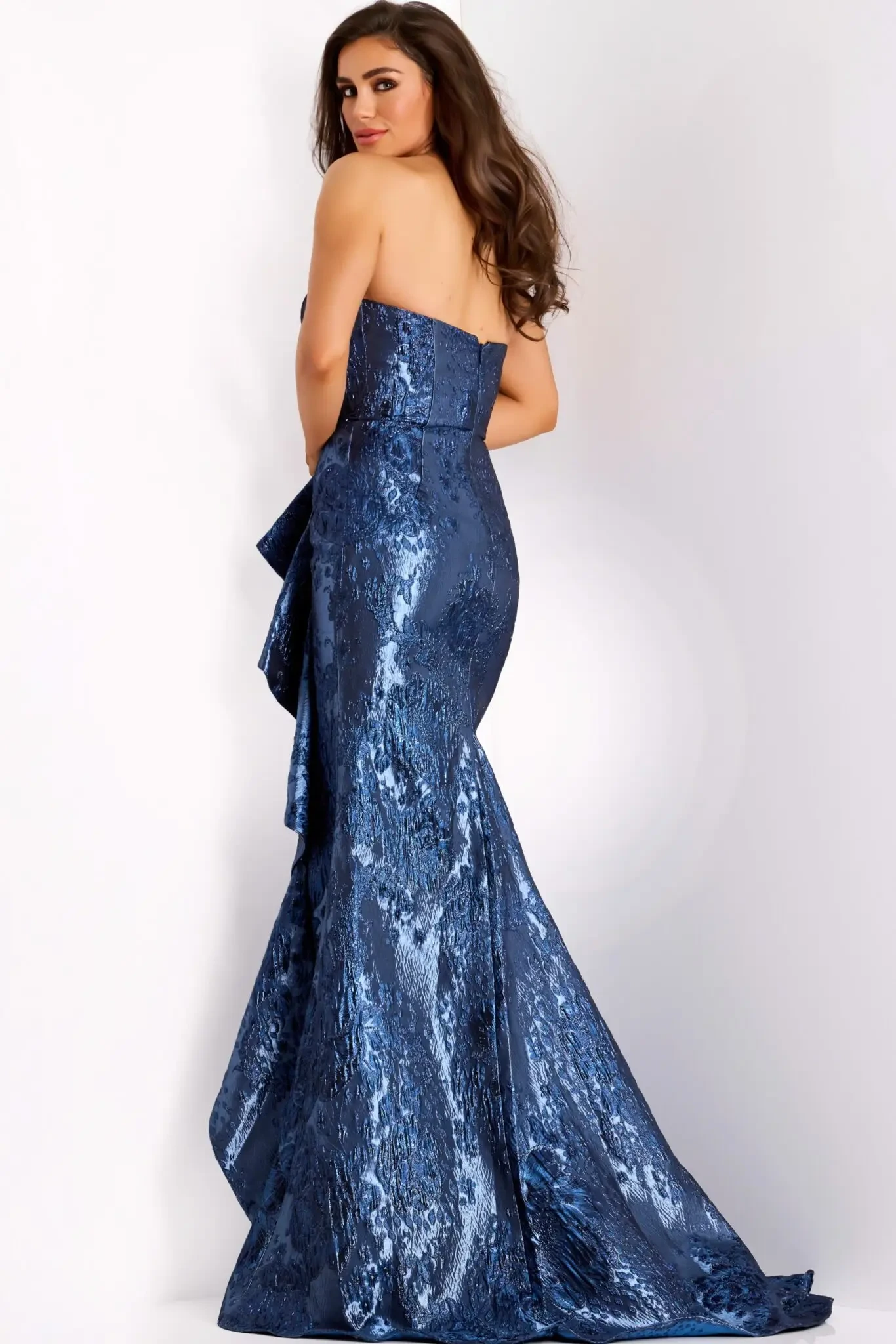 Model is wearing a strapless mermaid gown with side ruffles, fitted bodice and hips, a flared knee-length skirt, and metallic pleated detailing