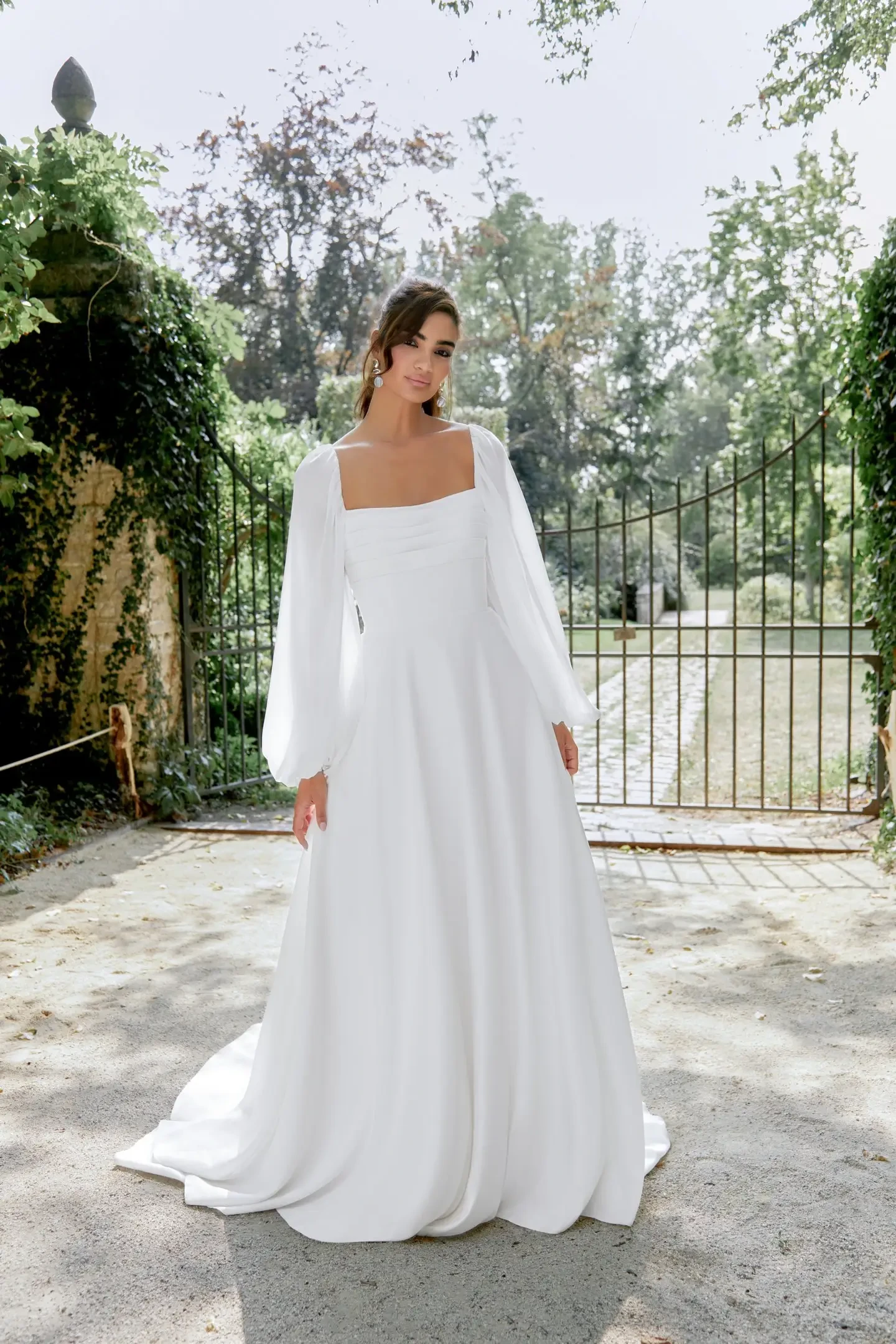 Model is wearing a satin A-line wedding gown with a pleated bodice, chiffon blouson sleeves, and a flowing skirt