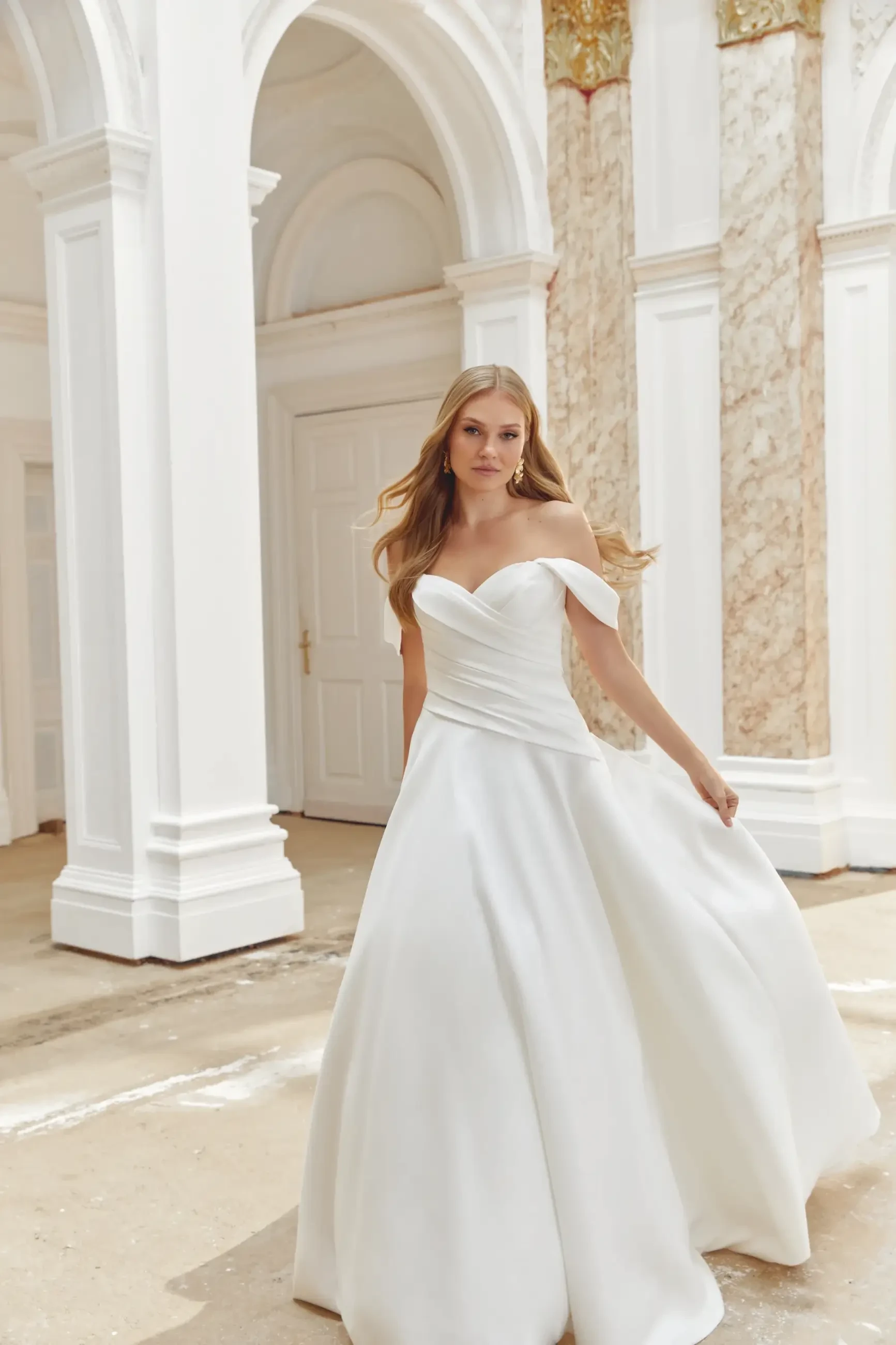 A woman in an elegant, off-shoulder white wedding gown stands in a grand, ornate setting with marble columns and arched doorways, exuding grace.
