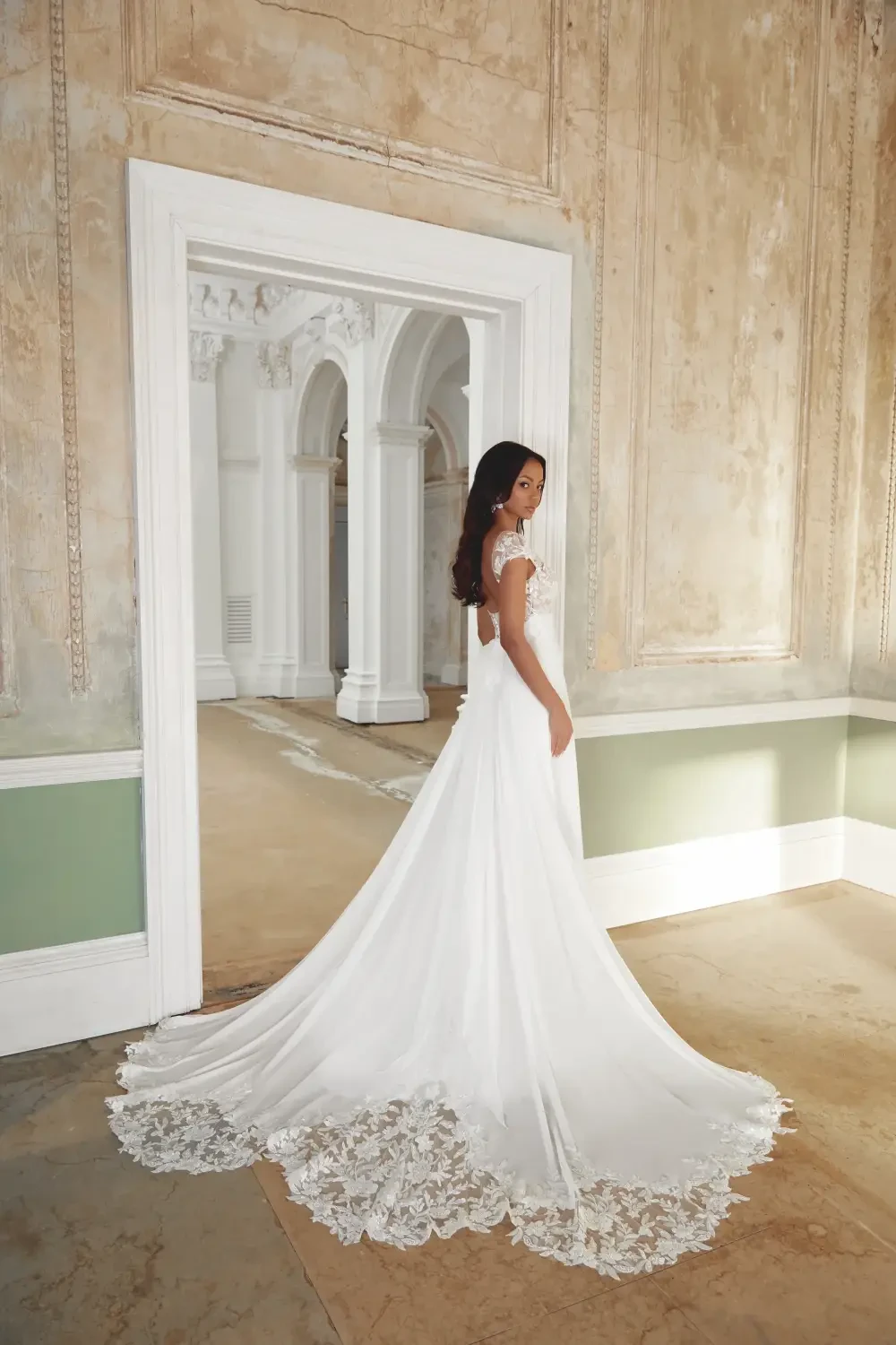 A woman wearing a white wedding dress standing in a doorway, showcasing the back of the dress and its lace details.