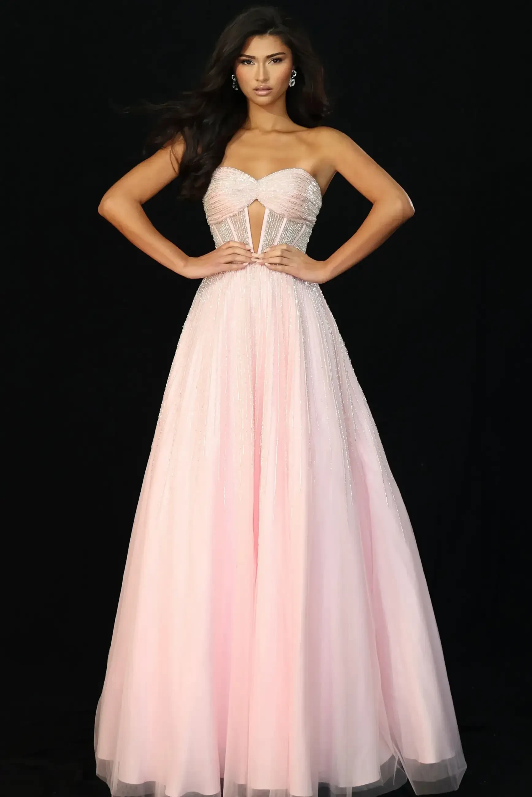 A model wearing a strapless, light pink gown with a fitted bodice and flowing skirt, posing against a black background.