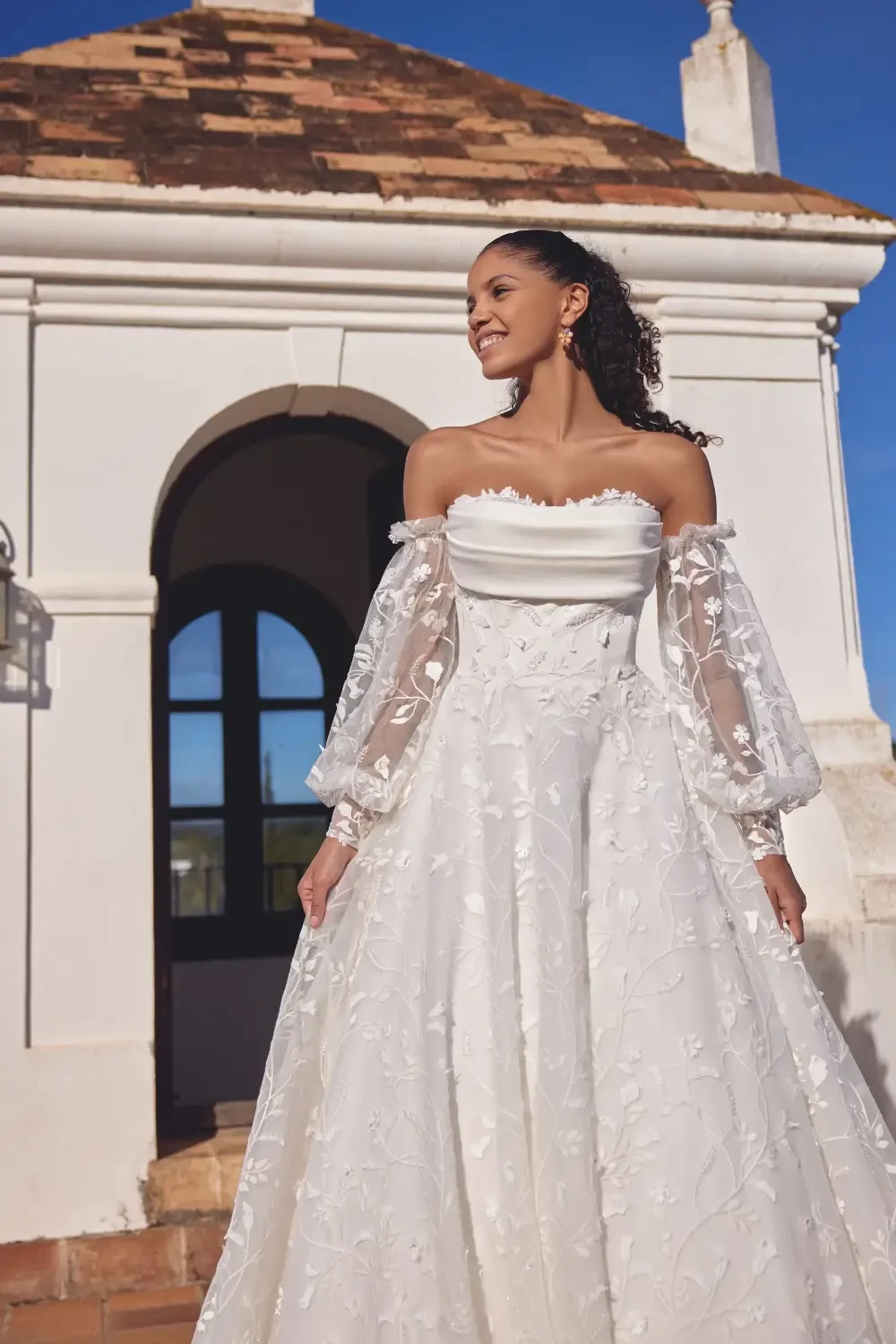 A woman in a white dress with off-the-shoulder sleeves and floral embroidery stands outside a building with a tiled roof, smiling.