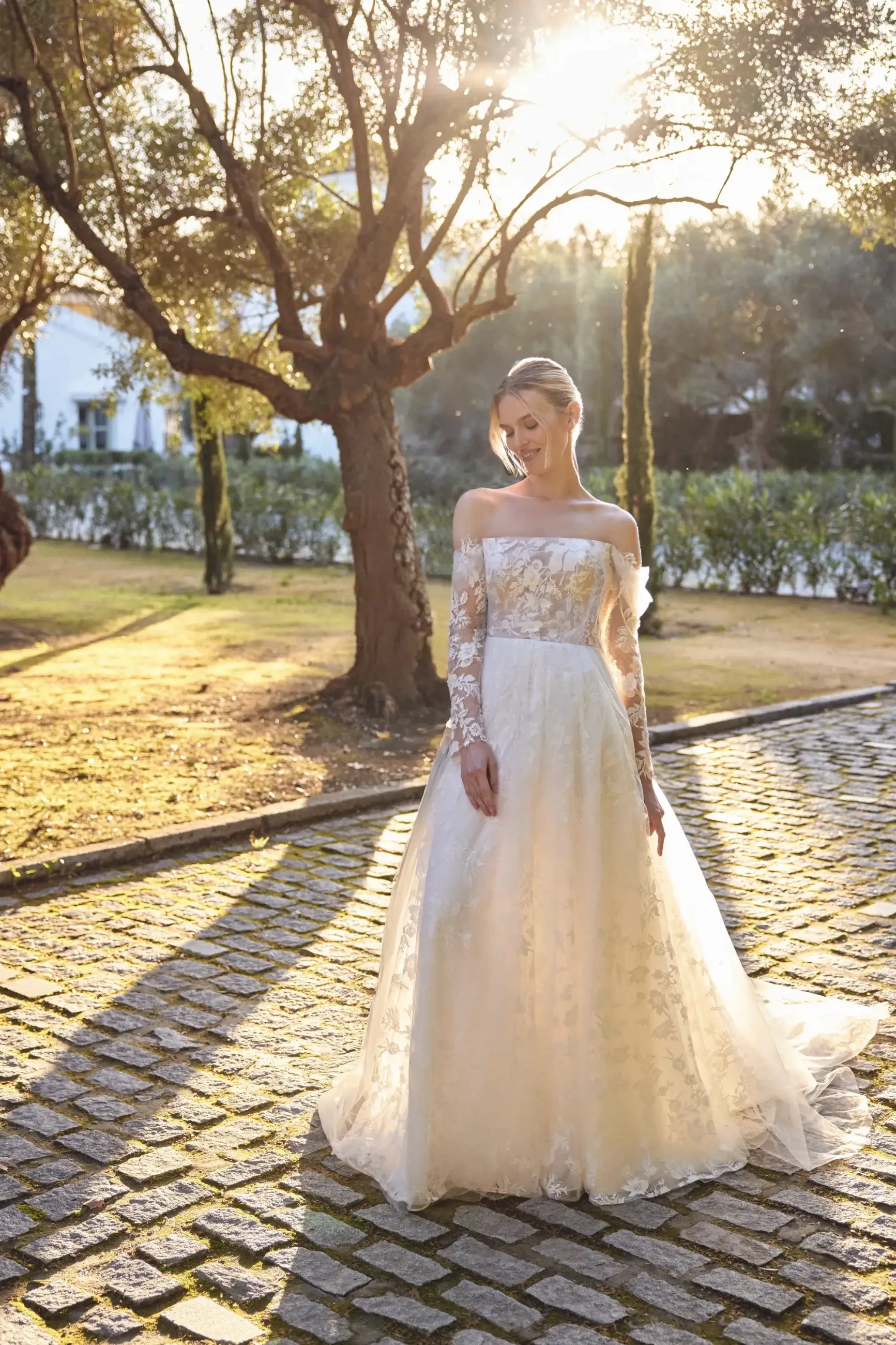 Bride in a delicate lace gown stands on a sunlit cobblestone path, surrounded by trees. The scene radiates warmth and serenity.