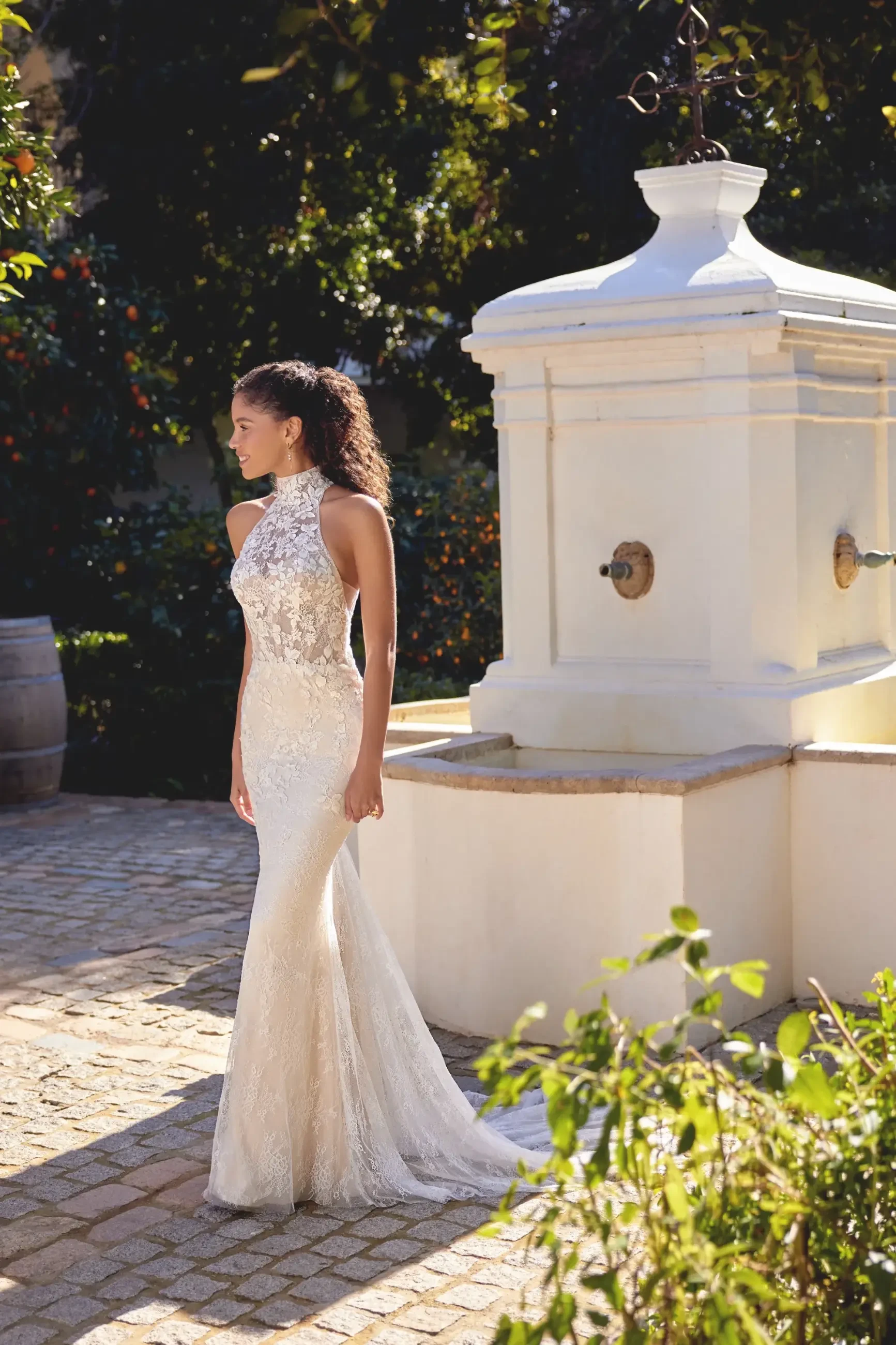 Bride in an elegant, lace mermaid wedding gown stands by a white stone fountain in a sunlit garden, exuding a serene and romantic ambiance.