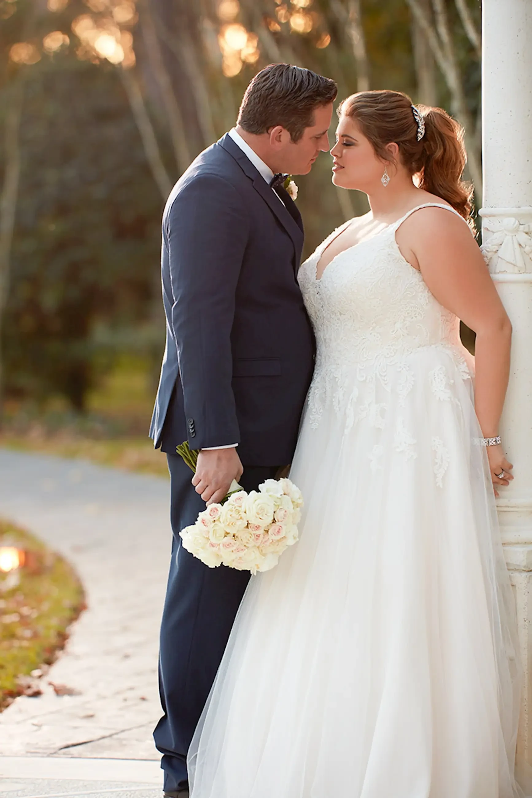 A couple in formal wedding attire sharing a romantic moment outdoors. The bride is holding a bouquet of white flowers.