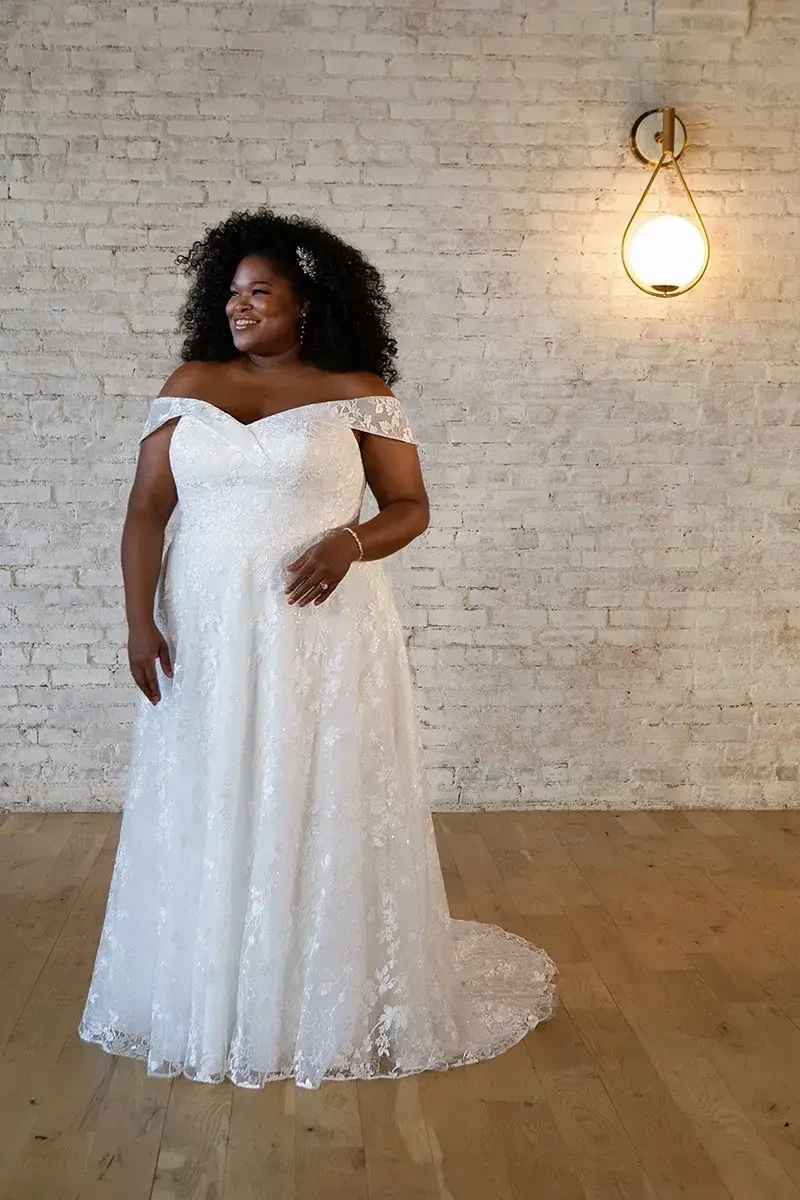 A woman with curly hair is wearing a white off-the-shoulder wedding dress, standing in a well-lit room with a brick wall and an exposed light fixture.