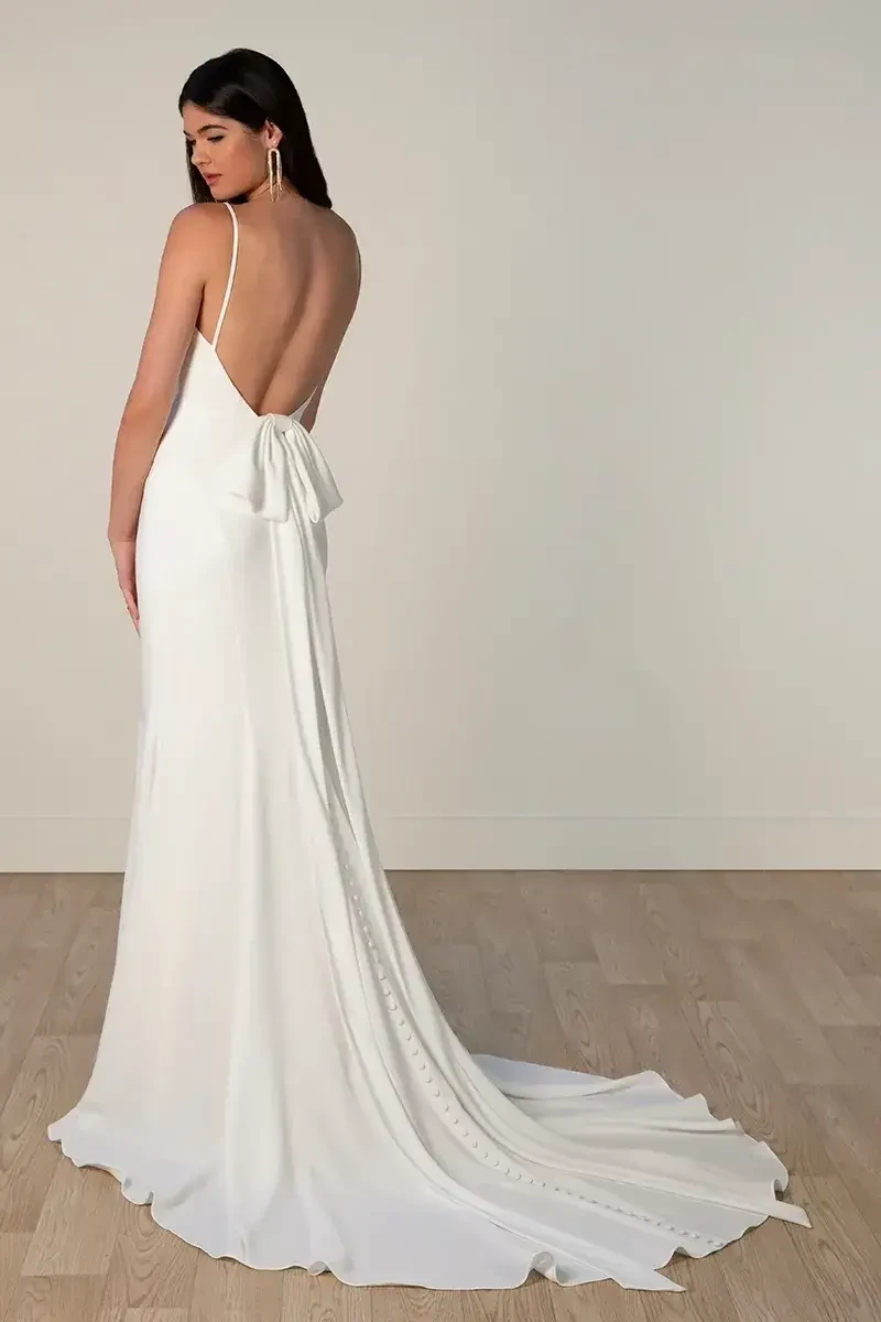 Model is wearing a sleek, modern crepe column wedding gown with a scoop neckline, spaghetti straps, low open back, and a detachable oversized bow.