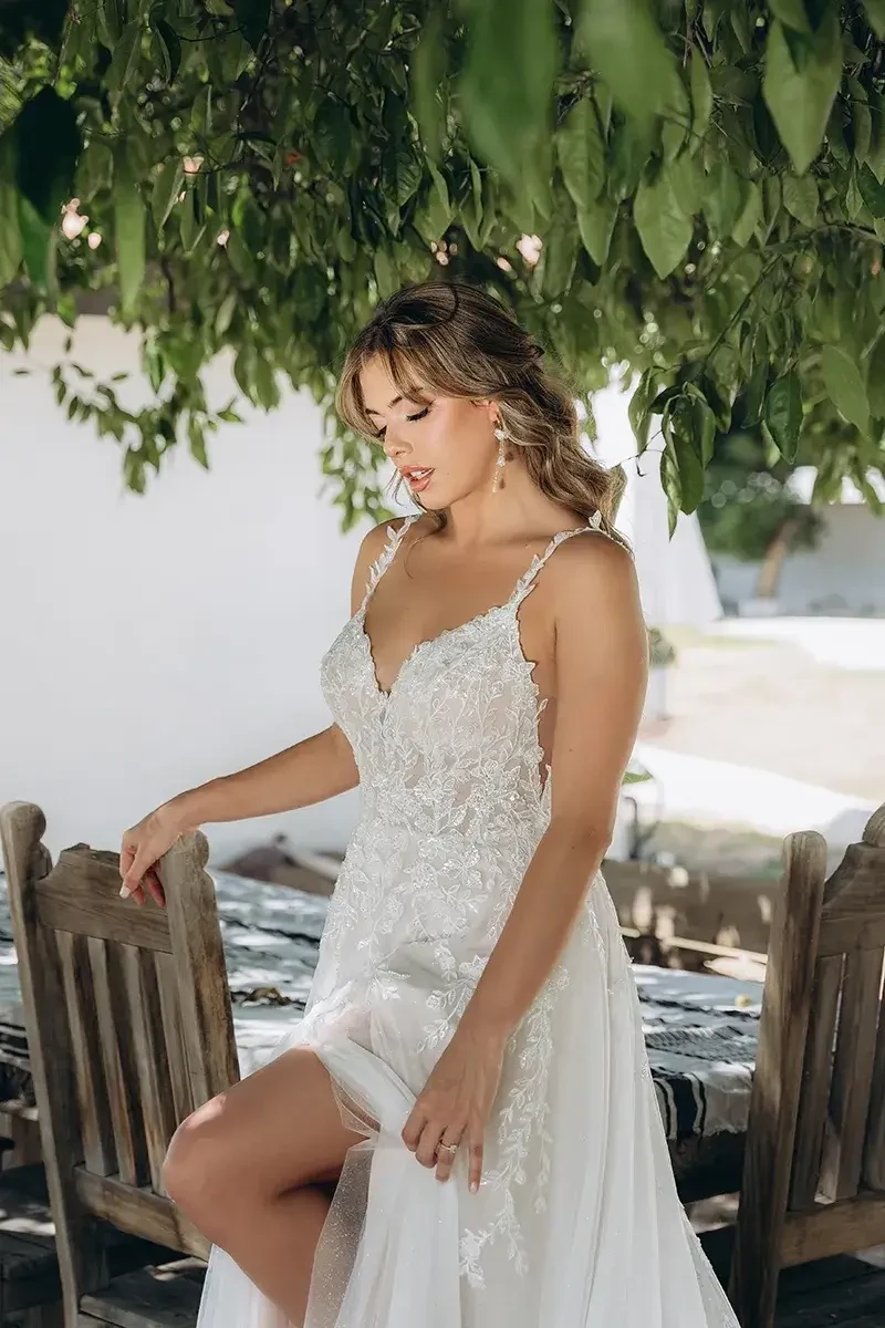 Model is wearing an ethereal A-line gown with shimmer tulle, lace appliqués, a sheer bodice with side cutouts, keyhole back, and a front slit 