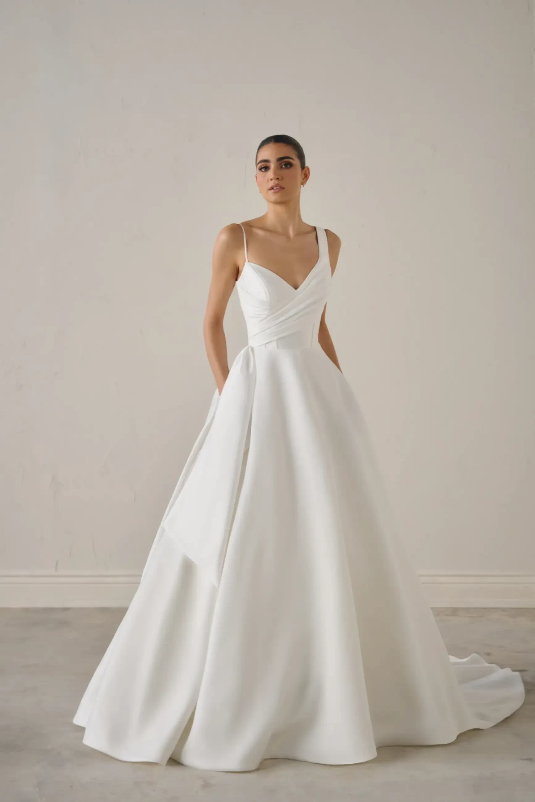 A woman in an elegant, flowing white bridal gown stands against a plain light gray background. The dress features thin straps and a fitted bodice, conveying a modern, sophisticated tone.