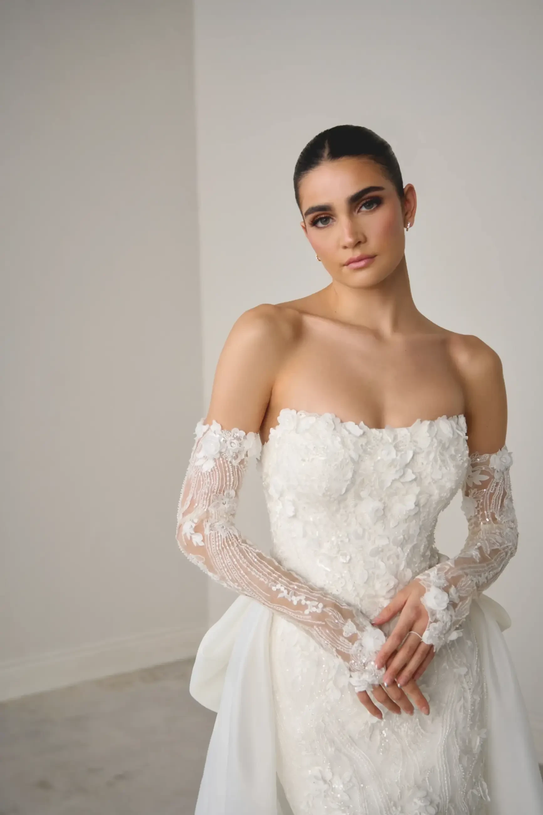 Bride in an elegant, off-shoulder lace wedding gown with intricate floral details, standing against a plain backdrop, exuding a serene, graceful aura.