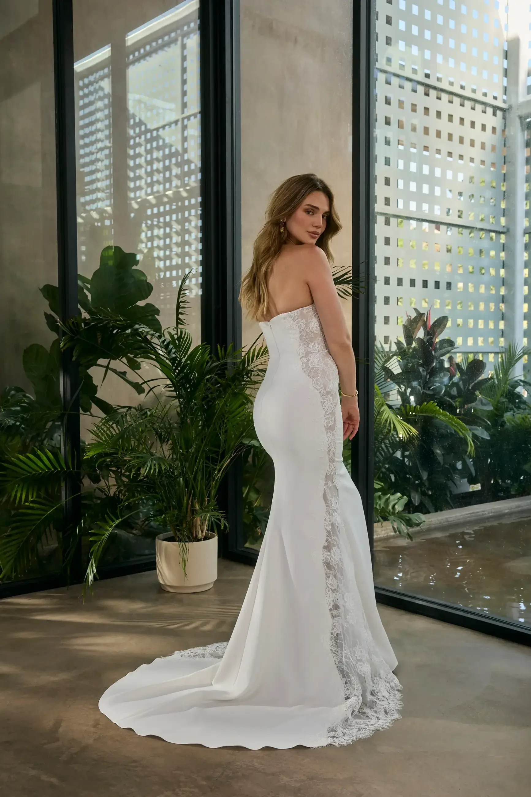A woman in a strapless, white lace wedding gown stands gracefully in a modern space with large windows and lush indoor plants, exuding elegance.