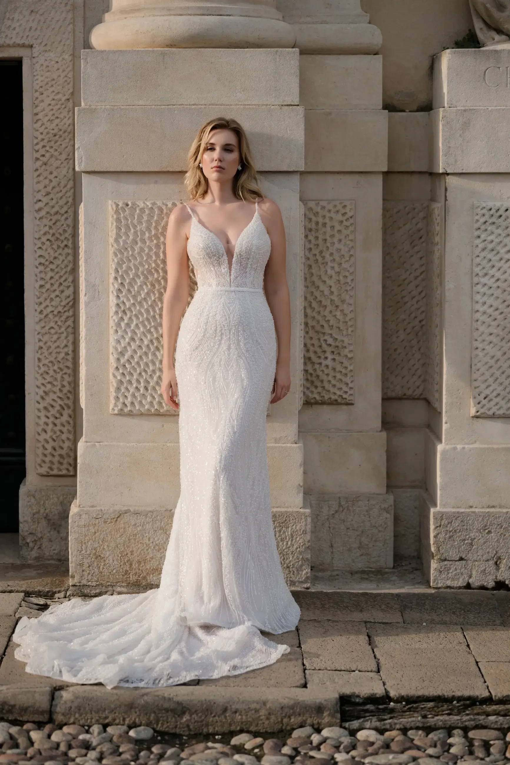 Model is wearing a sleeveless sheath wedding gown adorned with beaded lace appliques, a plunging neckline, and lightweight fabric