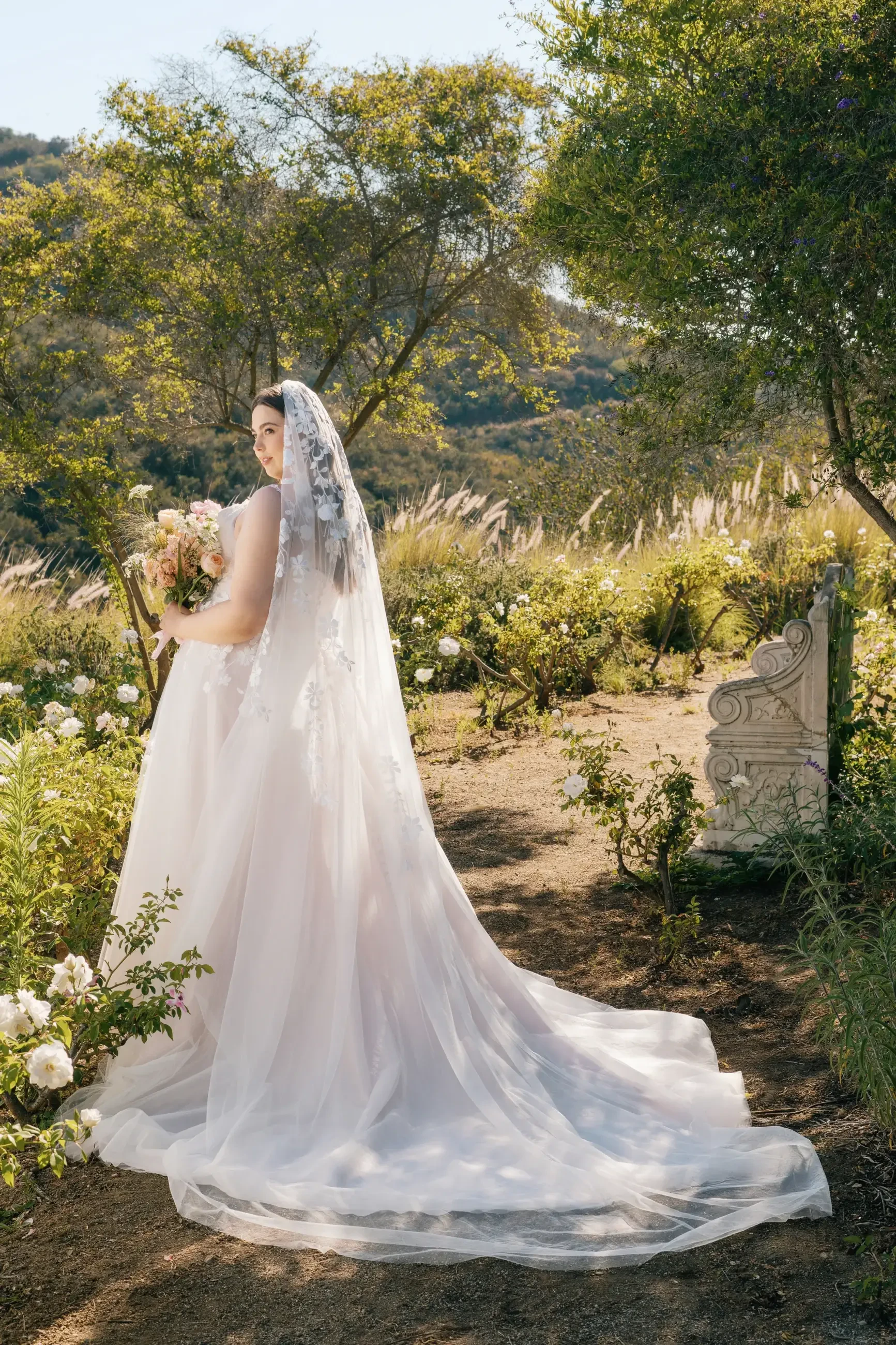 A bride in a flowing white gown and veil stands in a lush garden, holding a bouquet of flowers, surrounded by greenery and blooming plants.
